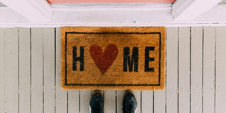 A pair of feet near a doormat with the word Home written on it, the letter o is a heart