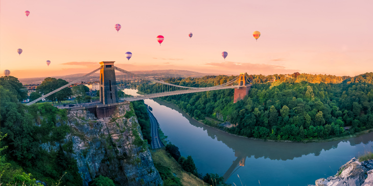 A bridge over a river with hot air balloons floating in the background on a sunset