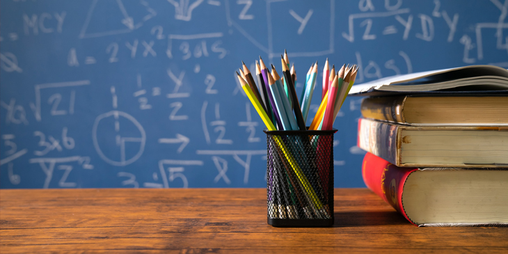 A pile of book and a collection of pencils in a pencil cup on a table, with a blackboard filled with mathematics in the background